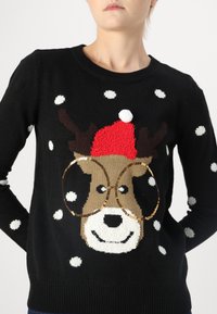 Pull noir avec un visage de renne portant un chapeau rouge et de grandes lunettes à sequins dorés, agrémenté de motifs de flocons de neige blancs.