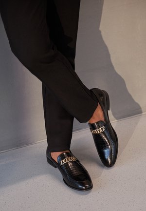 Sorte loafers lavet af krokodilembosseret læder, med en guldkæde over vristen og en glat, blank finish.