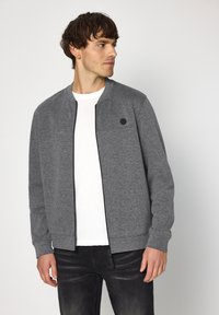 Grijze bomberjack met ritssluiting aan de voorkant, geribbelde manchetten en een klein zwart logo op de borst; gedragen over een wit t-shirt en donkere jeans.