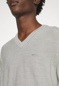 Gros plan d’une personne portant un pull gris clair à col en V avec le mot « BOSS » brodé sur la poitrine.