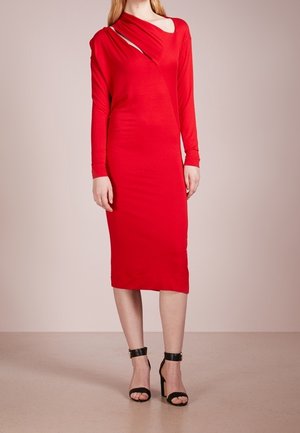Vrouw draagt een rode midi-jurk met lange mouwen en asymmetrische halsuitsnijdingen, gecombineerd met zwarte enkelsandalen met hoge hak en enkelriempje.