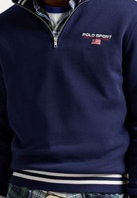 Navy zip-up sweatshirt with white striped collar and cuffs, featuring "Polo Sport Ralph Lauren" logo and American flag emblem.