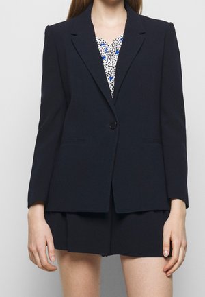 Mujer con blazer azul marino con un solo botón sobre una blusa estampada y shorts azul marino a juego sobre un fondo liso.
