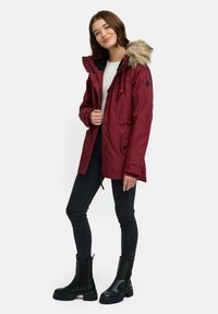 Giacca parka color bordeaux con cappuccio foderato in pelliccia, dotata di cintura regolabile in vita, tasche laterali e un badge nero sulla manica sinistra. Abbinata a jeans skinny neri e stivali neri robusti.