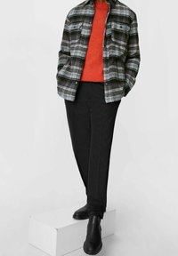 Rutig svart och grå flanellskjorta över en orange tröja, kombinerad med svarta byxor och svarta ankelboots, stående på en vit plattform.