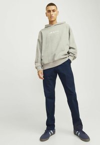 Ung mand står med den ene hånd i lommen, iført en lysegrå hættetrøje, mørkeblå bukser og marineblå sneakers med hvide striber.