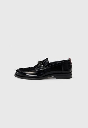 Black patent leather loafers with a low heel, decorative stitching, and a silver-tone buckle. Features a contrasting red pull tab at the back.
