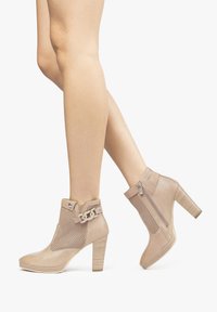 Stivaletti beige alla caviglia con design perforato, tacco a blocco chunky, zip laterale e accentuazione a catena decorativa sulla parte superiore. Texture liscia, forma moderna.