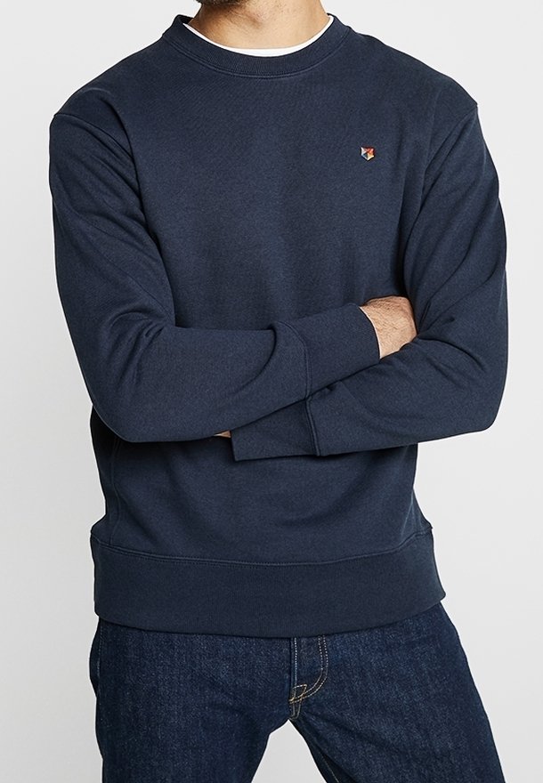 Homme les bras croisés portant un sweat-shirt bleu marine foncé et un jean bleu, affichant un petit logo géométrique multicolore sur la poitrine.