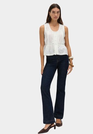Vrouw draagt een mouwloze witte blouse met kanten rand, donkerblauwe rechte jeans, bruine slingbackhakken en armbanden aan de rechterpols.