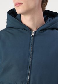 Chaqueta con capucha en un verde azulado oscuro, hecha de una tela texturizada. Presenta un zipper plateado y un cuello alto, con un diseño limpio y moderno.