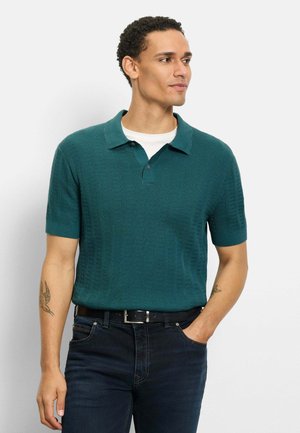 Mann trägt ein türkisfarbenes kurzärmeliges Strick-Poloshirt, das in dunkle Jeans gesteckt ist, steht mit der rechten Hand in der Tasche und schaut zur Seite.