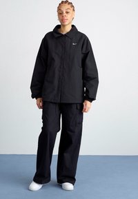 Juodas neperšlampamas švarkas su apykakle ir priekinėmis kišenėmis, derinamas su laisvais krovininiais džinsais. Viršutiniame kairiajame kampe yra maža balta logotipas.