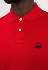 Camiseta polo roja hecha de tela texturizada, con cuello clásico, tres botones blancos y un logo bordado en azul en el lado izquierdo del pecho.
