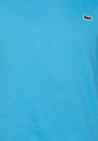 Maglietta in cotone azzurro chiaro con una texture liscia, caratterizzata da un piccolo logo di un coccodrillo verde ricamato sul davanti.