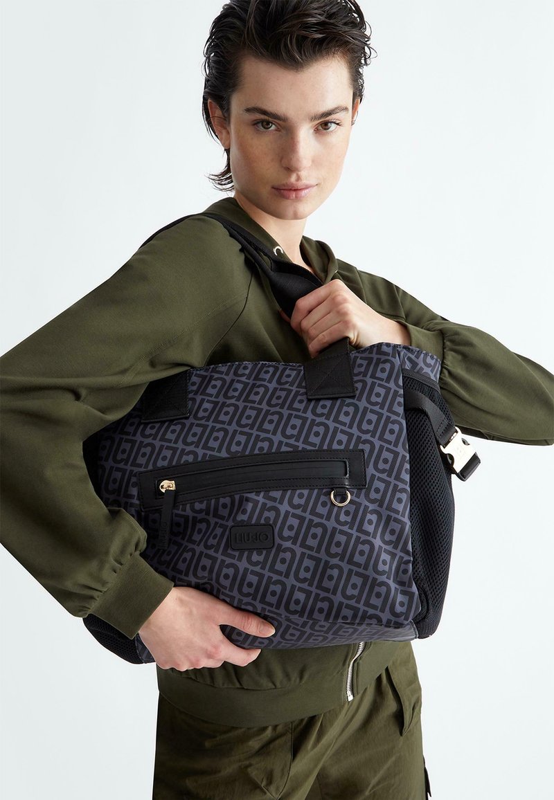 Borsa a tracolla nera e grigia con pattern geometrico, dotata di tasca frontale con zip, manici robusti e dettagli in rete. Il materiale sembra resistente.