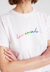 Bílý bavlněný tričko s barevným nápisem "láska znamená láska" ve stupních šedi. Krátké rukávy a kulatý výstřih.