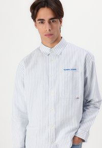 Joven vestido con una camisa de botones a rayas en azul claro y blanco, con "TOMMY JEANS" bordado sobre el bolsillo del pecho, con la mano en el bolsillo.