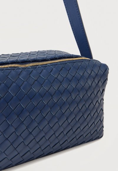 Μπλε τσάντα χιαστί με υφή υφασμένου δέρματος και λεπτομέρεια χρυσού φερμουάρ σε απλό ανοιχτό φόντο.