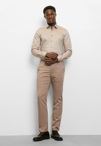 Eng anliegendes beiges Hemd mit Knöpfen und einem dezenten Muster, kombiniert mit passenden beigen Hosen und dunkelbraunen eleganten Schuhen.