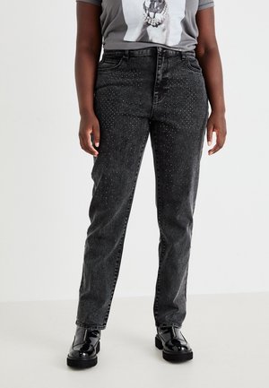 Osoba nosząca ciemnoszare dżinsy z rozproszonymi małymi kryształkami, w połączeniu z szarą, graficzną koszulką i czarnymi błyszczącymi butami.