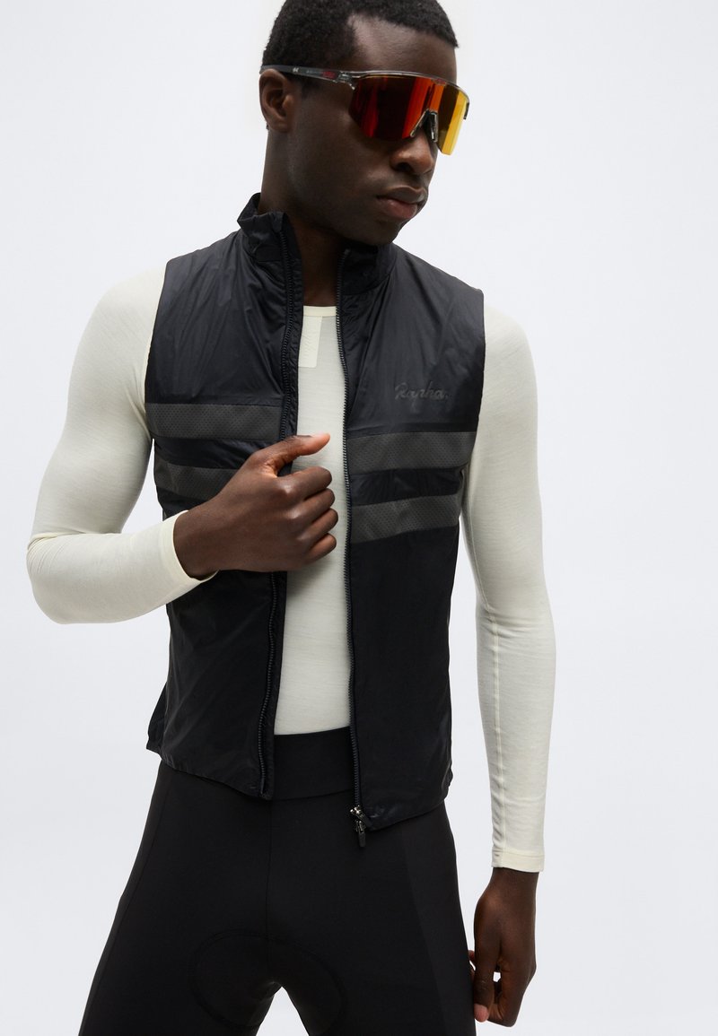 Man wearing black zip-up cycling vest over a long-sleeve white base layer and red-tinted sports sunglasses, adjusting the vest.