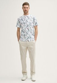 Camiseta blanca de manga corta con un patrón de hojas verdes y rosas, combinada con pantalones beige claros y zapatillas blancas. Atuendo simple y casual.