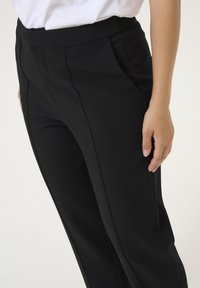 Pantaloni neri con una texture liscia, design strutturato, vita larga e pieghe sottili. Mano appoggiata delicatamente sulla coscia.