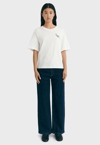 Weißes Baumwoll-T-Shirt mit kurzen Ärmeln, das mit einem kleinen Blumenmuster versehen ist. Kombiniert mit lockeren dunkelblauen Jeans. Schwarze Schuhe.