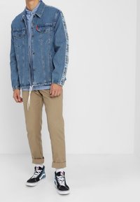 Homme portant une veste en denim bleu avec bande latérale, une chemise boutonnée bleu clair, un pantalon beige retroussé et des baskets montantes noires avec des lacets blancs.