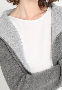 Grå ullcardigan med en ljusgrå huva, som har en mjuk textur. Under finnes en vit, åtsittande långärmad skjorta.