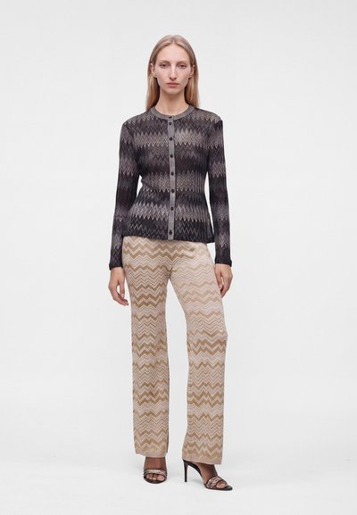 Femme debout portant un cardigan à boutons noir et gris en zigzag et un pantalon large beige à motif zigzag avec des sandales noires à talons hauts.