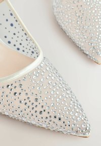Beige kengät, joissa on kärki, verkkomateriaali, ja koristeltu hopeisilla paljeteilla sekä satiinireunus aukossa, esittelee tekstuurin pinnan.