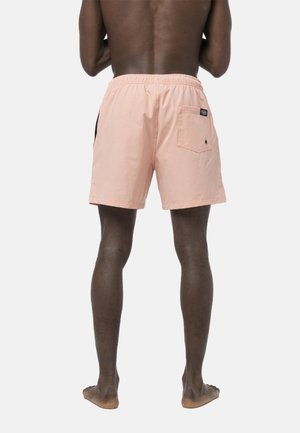 Homme debout, pieds nus, de dos, portant un short de bain couleur pêche avec une poche arrière et une ceinture élastique, sur un fond uni.