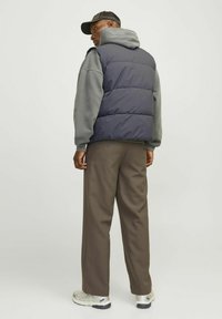 Padded námo vestu cez šedú mikinu, v kombinácii s hnedými nohavicami. Model je otočený k nám chrbtom, má na hlave šiltovku a svetlé tenisky.