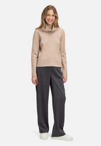 Eine lächelnde Frau trägt einen beigen, karierten Rollkragenpullover, lockere dunkelgraue Hose und weiße Sneakers, während sie vor einem einfarbigen Hintergrund steht.