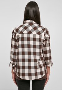 Femme aux longs cheveux foncés portant une chemise à carreaux marron et blanche à manches longues, debout, les bras détendus, dos au spectateur.