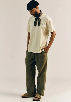 Mann mit schwarzer Baskenmütze, hellbeigem T-Shirt, olivgrüner Hose, schwarzem Halstuch und braunen Sandalen, steht mit den Händen in den Taschen.