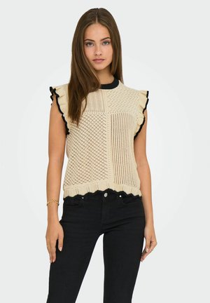 Top sans manches en beige avec des accents noirs, présentant un motif texturé et des bords volants. Porté avec un pantalon noir.