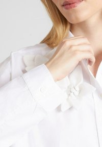 Camisa de cuello blanco con aplicaciones florales tridimensionales en blanco en el hombro. Textura de tela suave con detalle de botones en el puño.