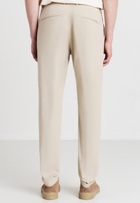 Personne portant un pantalon beige à jambes droites et des chaussures en daim tan, vue de dos sur un fond clair.