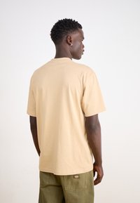 Un tricou din bumbac de culoare bej, cu gât rotund și mâneci scurte, expus din spate, asortat cu pantaloni cargo de culoare verde măsliniu.