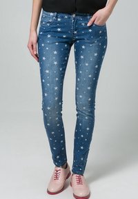 Jean en denim bleu avec des motifs d'étoiles blanches, coupe slim, design à cinq poches et légère décoloration. Associés à des chaussures à lacets rose pâle.