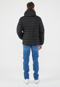 Giacca nera imbottita con cappuccio, caratterizzata da un design a cuciture verticali. Indossata con jeans blu e sneakers bianche, vista da dietro.