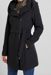 Mantello in misto lana grigio scuro con un ampio cappuccio, vita con cintura e chiusura frontale con zip. Presenta tasche laterali e una texture liscia.