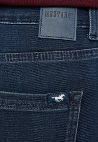 Sötétkék farmernadrág, amelyen egy "MUSTANG" feliratos bőrfolt és egy kis hímzett lólogó található a hátsó zseben.