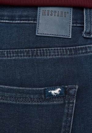 Jeans din denim albastru închis, cu o etichetă din piele inscripționată "MUSTANG" și un mic logo brodat cu un cal pe buzunarul din spate.