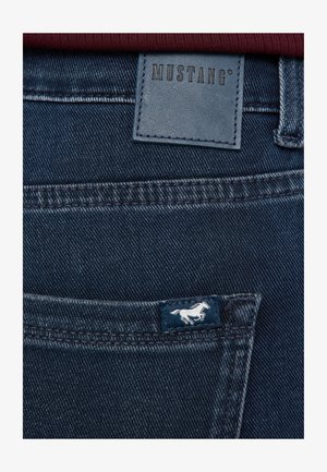 Jeans din denim albastru închis, cu o etichetă din piele inscripționată "MUSTANG" și un mic logo brodat cu un cal pe buzunarul din spate.