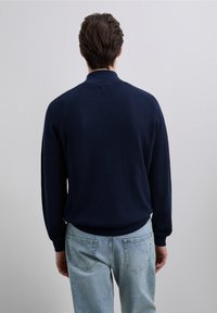 Donkerblauwe geribbelde trui met een staande kraag, lange mouwen en een ontspannen pasvorm, gecombineerd met lichtblauwe spijkerbroek.