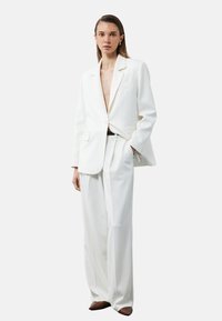 Weißes, maßgeschneidertes Blazer mit passenden weit geschnittenen Hosen. Glattes Material, tiefer V-Ausschnitt, Ein-Knopf-Verschluss, abgerundet mit einem schwarzen Gürtel.